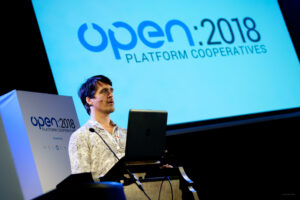 Oliver Sylvester-Bradley speaking at a conference with a laptop in front of a large screen that reads "OPEN 2018"