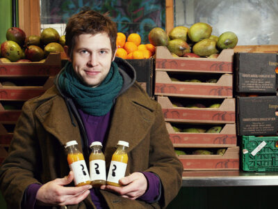Man wearing a scarf stands in front of wooden food bin holding three bottles of fresh made juices